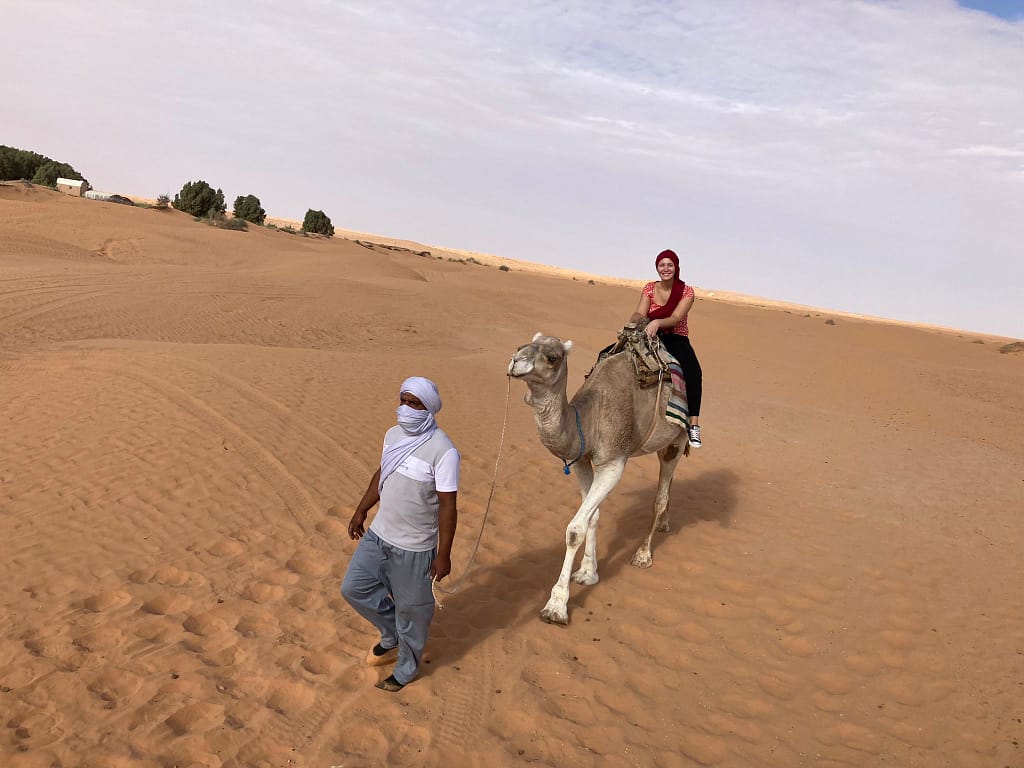 A desert guide leads a camel by a rope, with a tourist seated on its back, surrounded by the stunning dunes of the Tunisian desert.